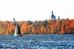 Un velero en un lago con follaje otoñal y un edificio al fondo.