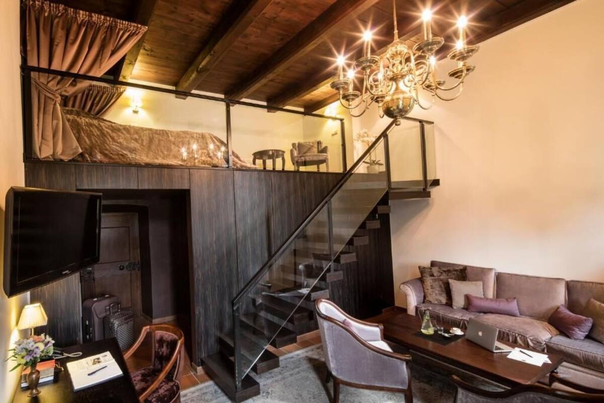 A modern loft-style room with a dark staircase, a flat-screen TV, a sofa, and a chandelier.