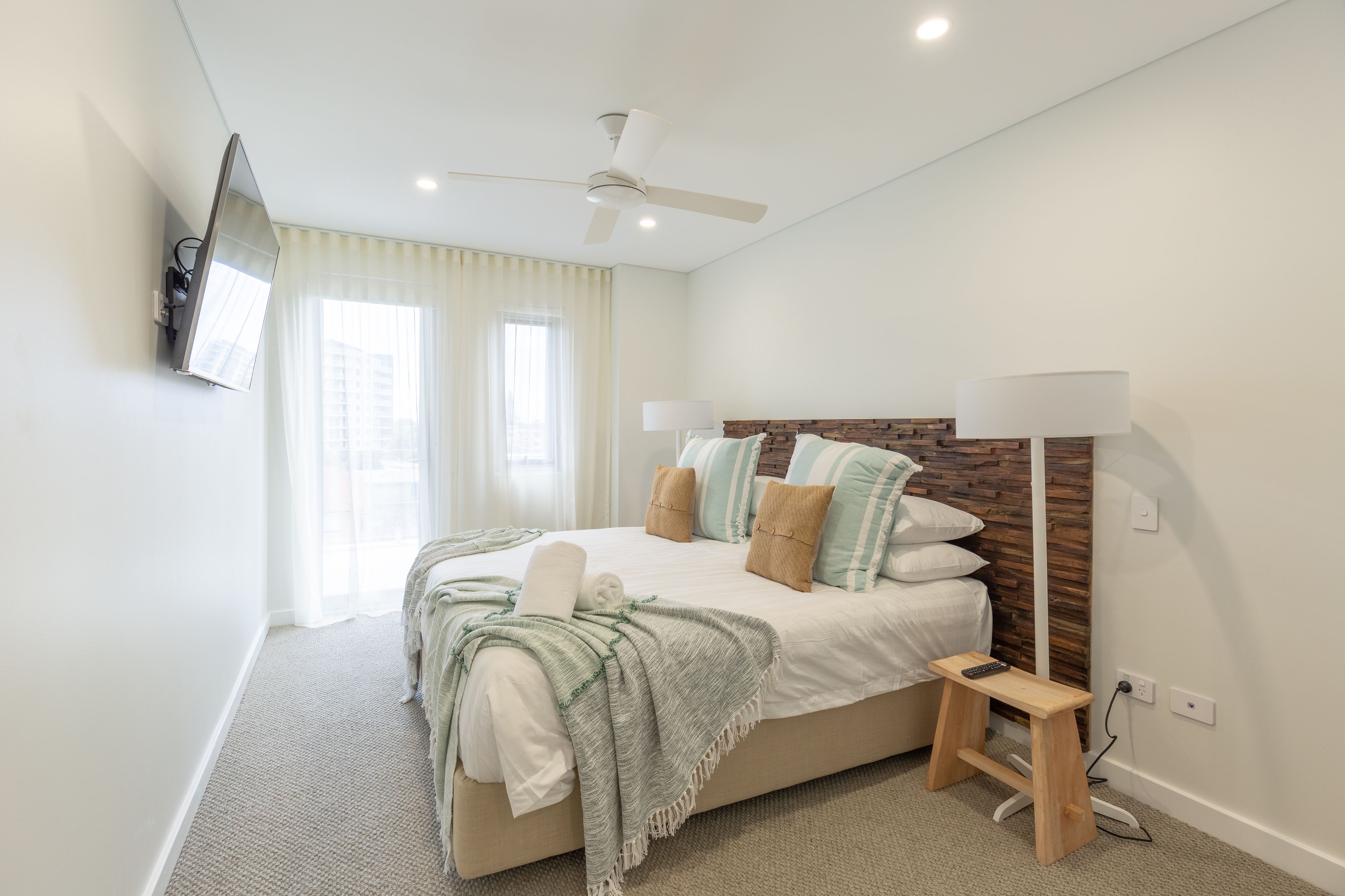 A modern bedroom with a bed, a standing lamp, a ceiling fan, and a TV mounted on the wall.