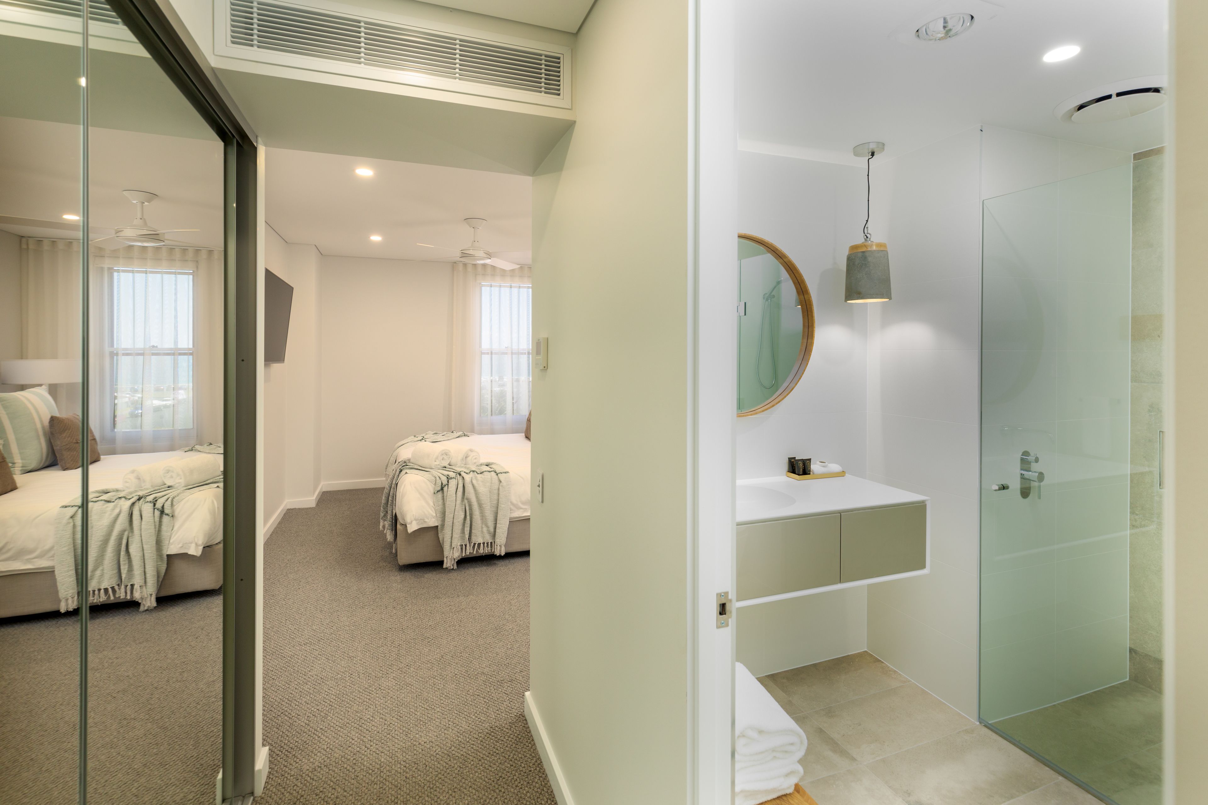 Una habitación de hotel moderna con cabina de ducha de vidrio, un espejo grande y una zona de dormitorio separada con cama y ventana.