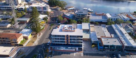 An aerial view of a coastal town with a prominent multi-story building with a flat roof, surrounded by smaller buildings and a marina.