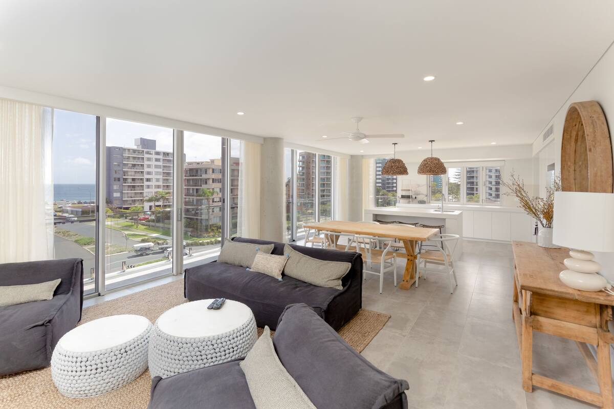 A modern living room with a large window, grey sofas, a wooden dining table, and a kitchen area.