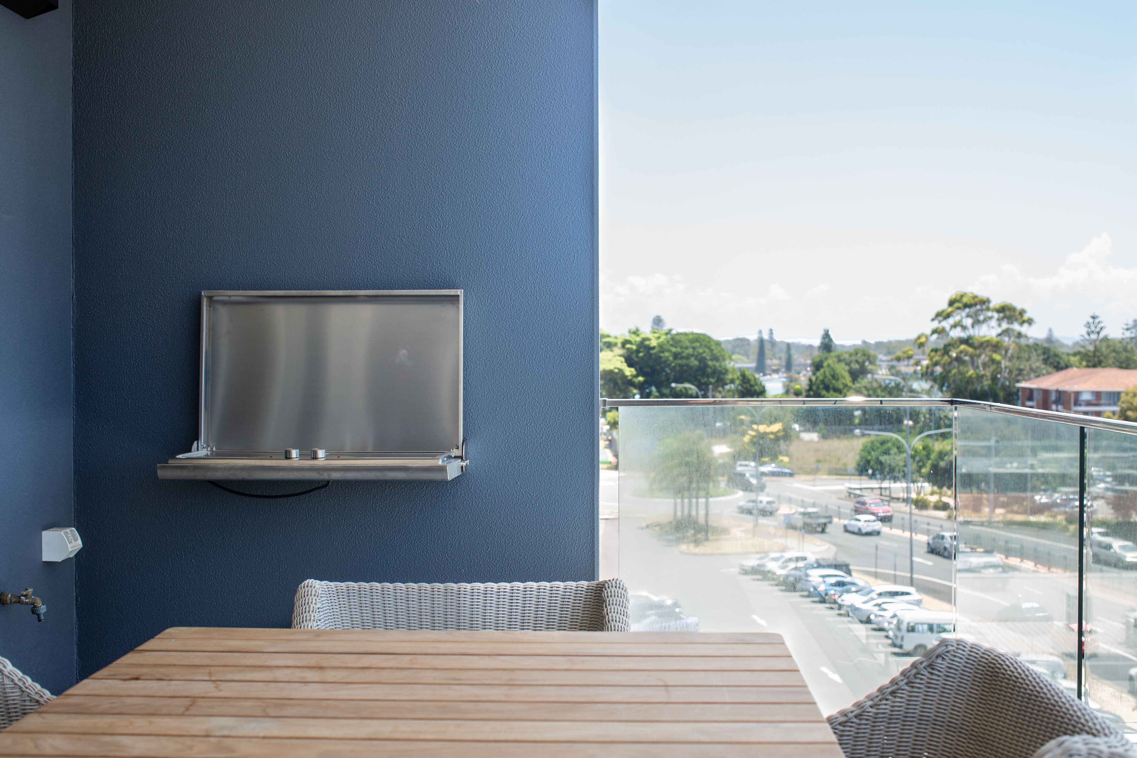 A balcony with a TV mounted on the wall, wicker furniture, and a view of a parking lot and street.