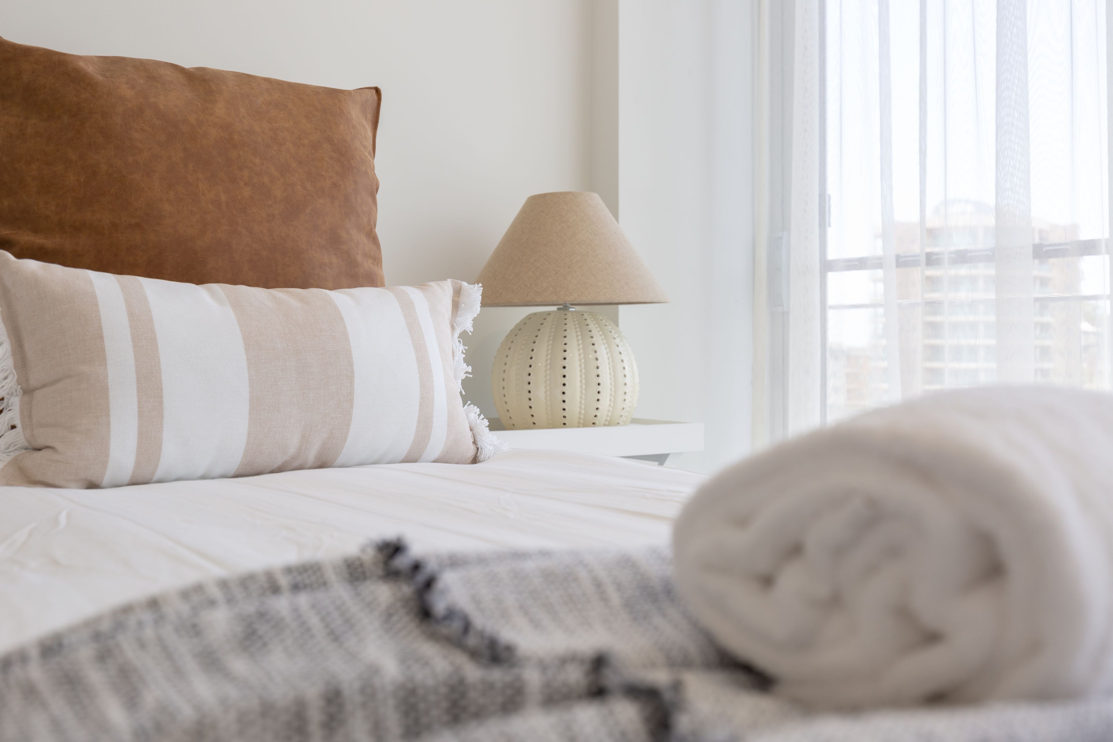A neatly made bed with striped pillows, a rolled white towel, and a beige lamp on a bedside table.