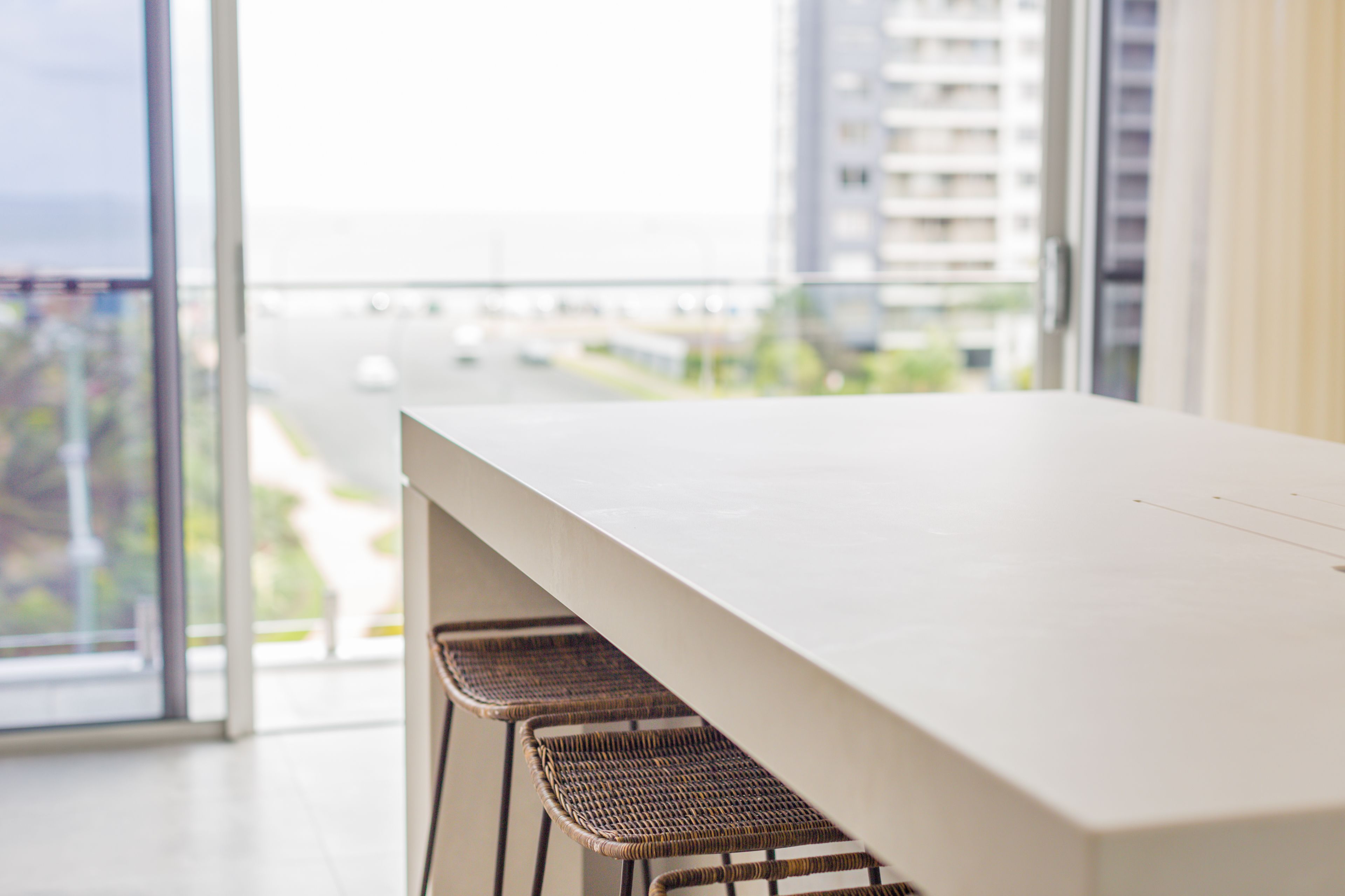 A modern interior with a white table and wicker chairs, large windows offering a view of a cityscape and a body of water.