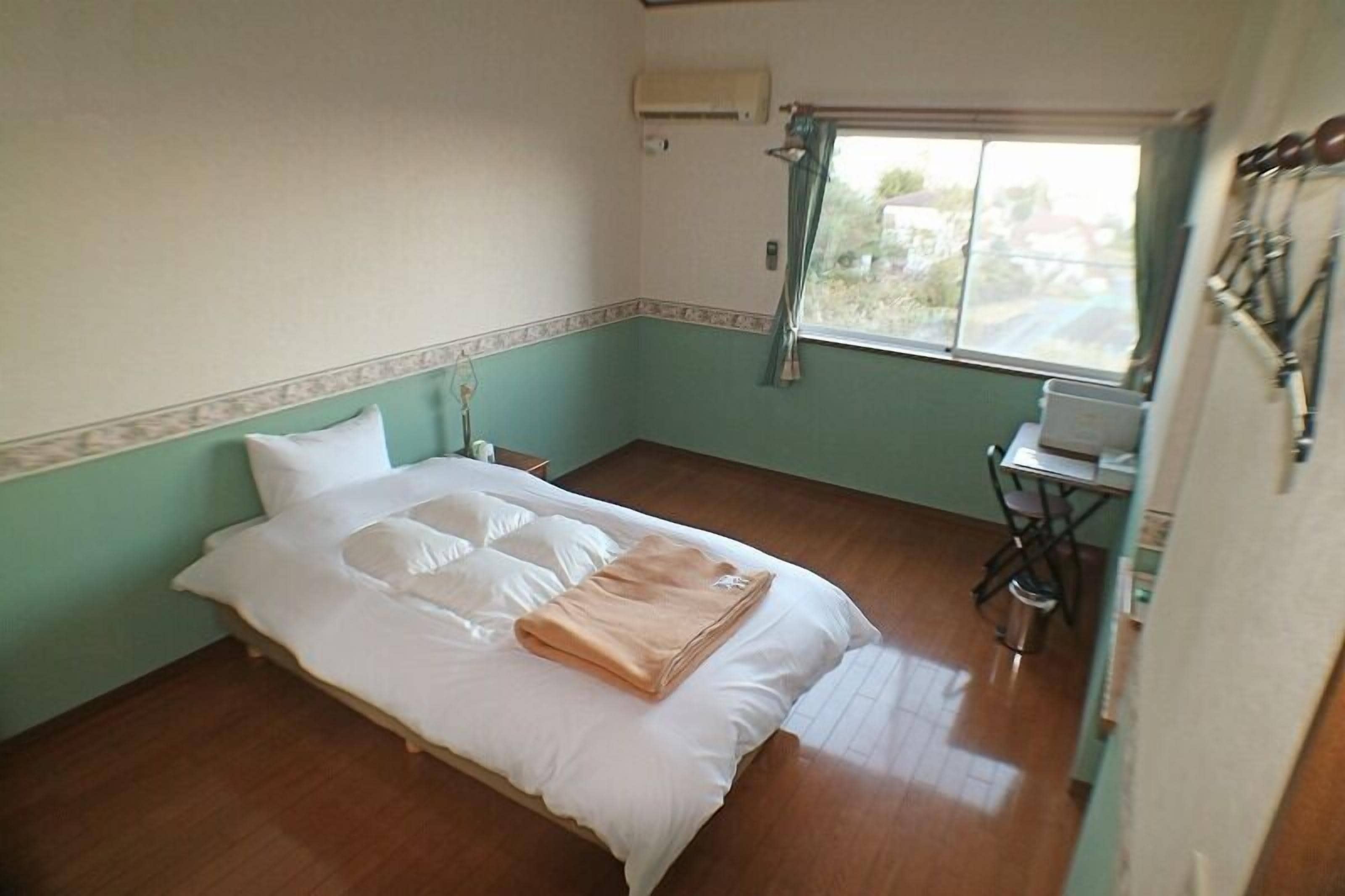 A small, single-bed room with a wooden floor, a window with curtains, a desk with a chair, and an air conditioning unit on the wall.