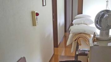 A hotel room with a bed, a bedside table with a remote control, a suitcase, a small table with a fan, and a water dispenser.