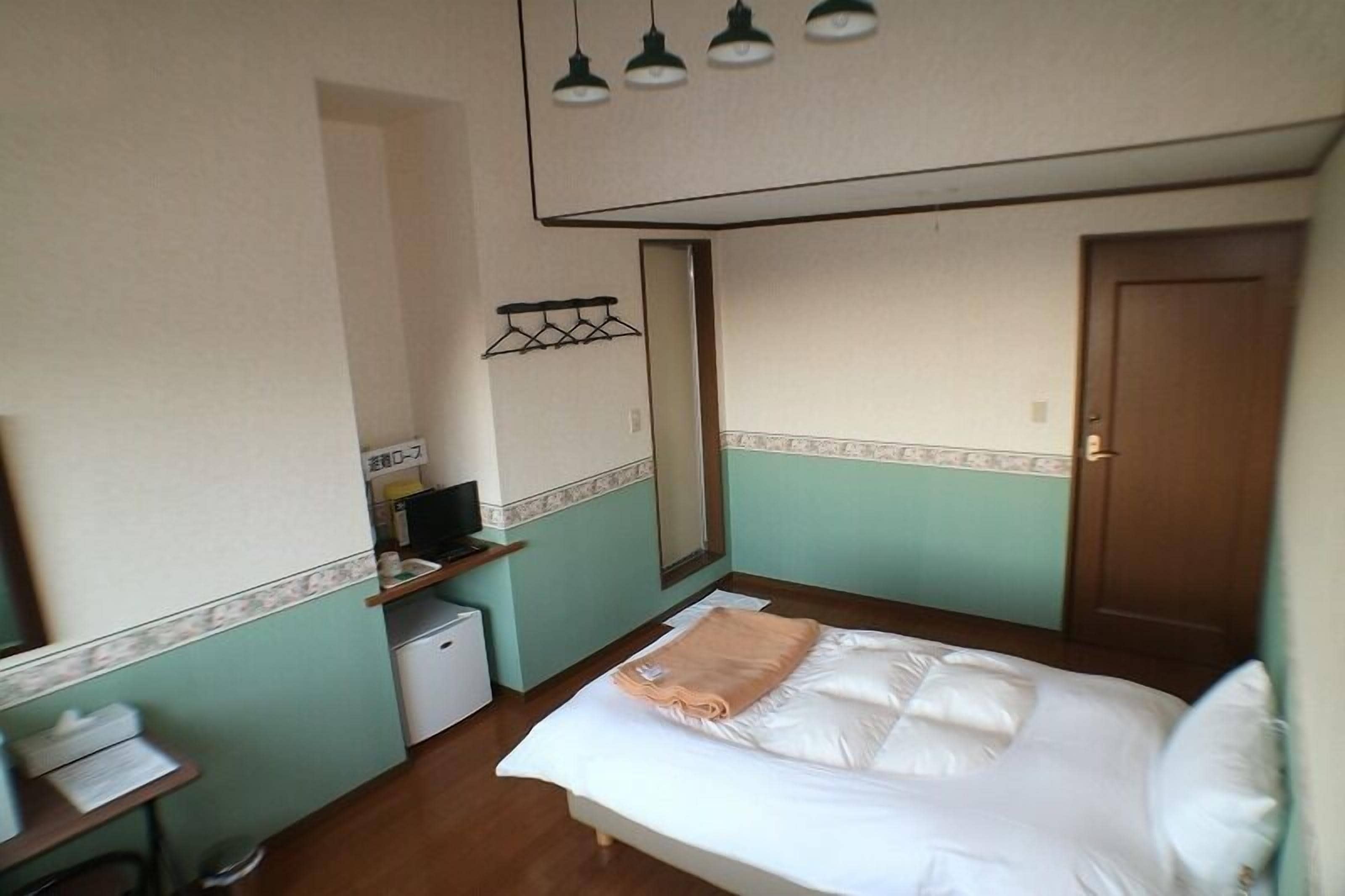 A small, compact hotel room with a single bed, a desk with a computer, a mirror, and a wardrobe.