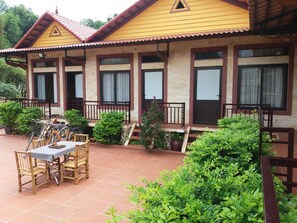 A yellow building with a tiled patio, wooden furniture, and potted plants. - Mai Chau Xanh Bungalow (Mai Chau)