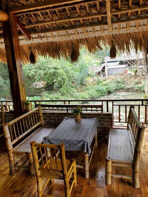 Área de estar ao ar livre com estrutura de bambu, mesa e cadeiras, com vista para um jardim e um pequeno riacho.
