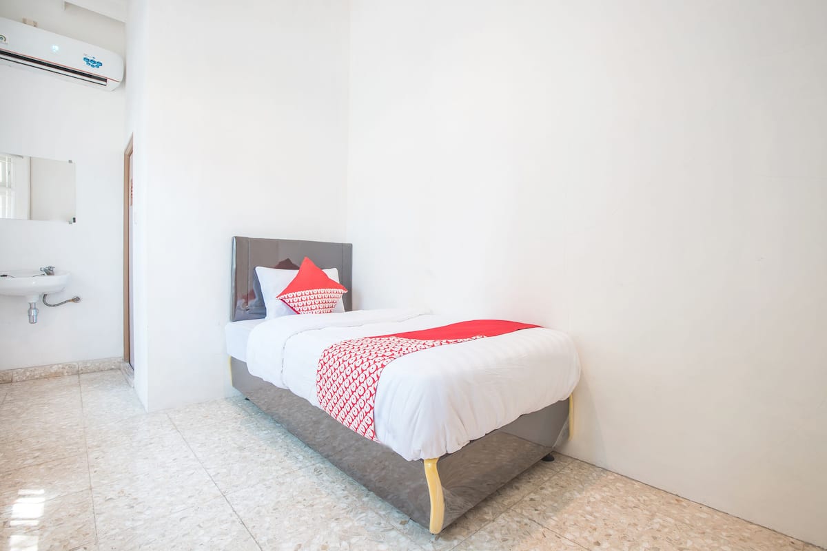 Une chambre à coucher propre et moderne, avec un lit, un coussin aux motifs rouges et blancs, un mur blanc et une salle de bain équipée d’un lavabo et d’un miroir.