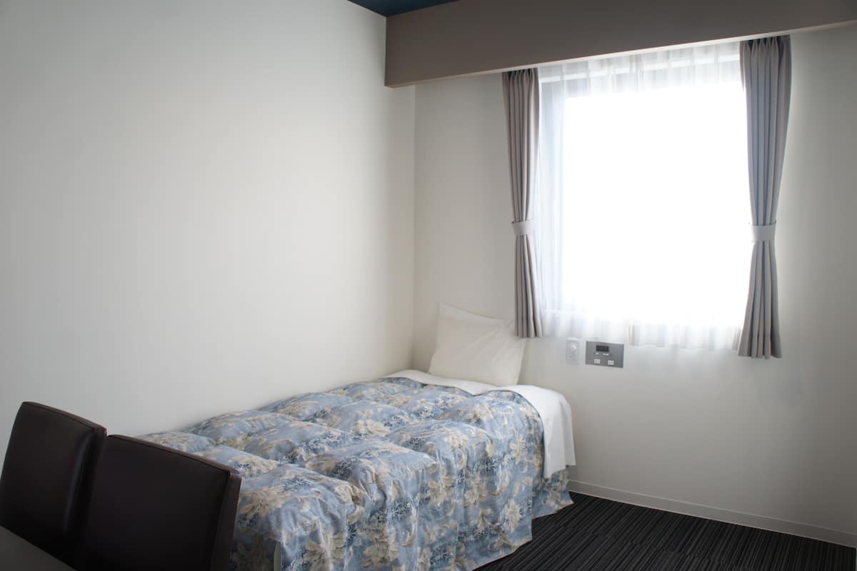 Een hotelkamer met een bed, een stoel, een raam met gordijnen en een thermostaat.