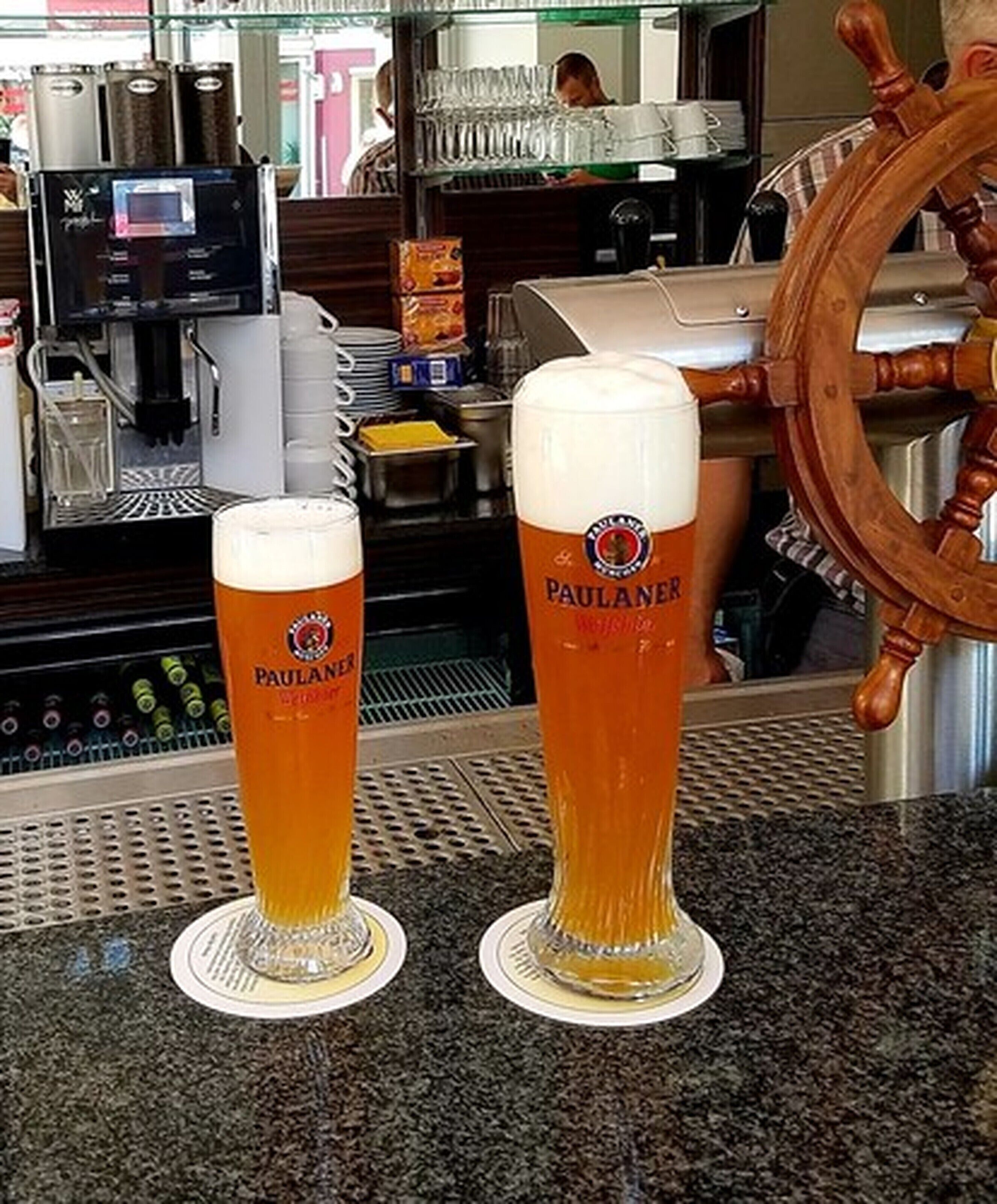 Two Paulaner beer glasses on a bar counter with a ship's wheel in the background.