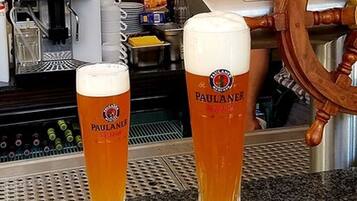 Two Paulaner beer glasses on a bar counter with a ship's wheel in the background.