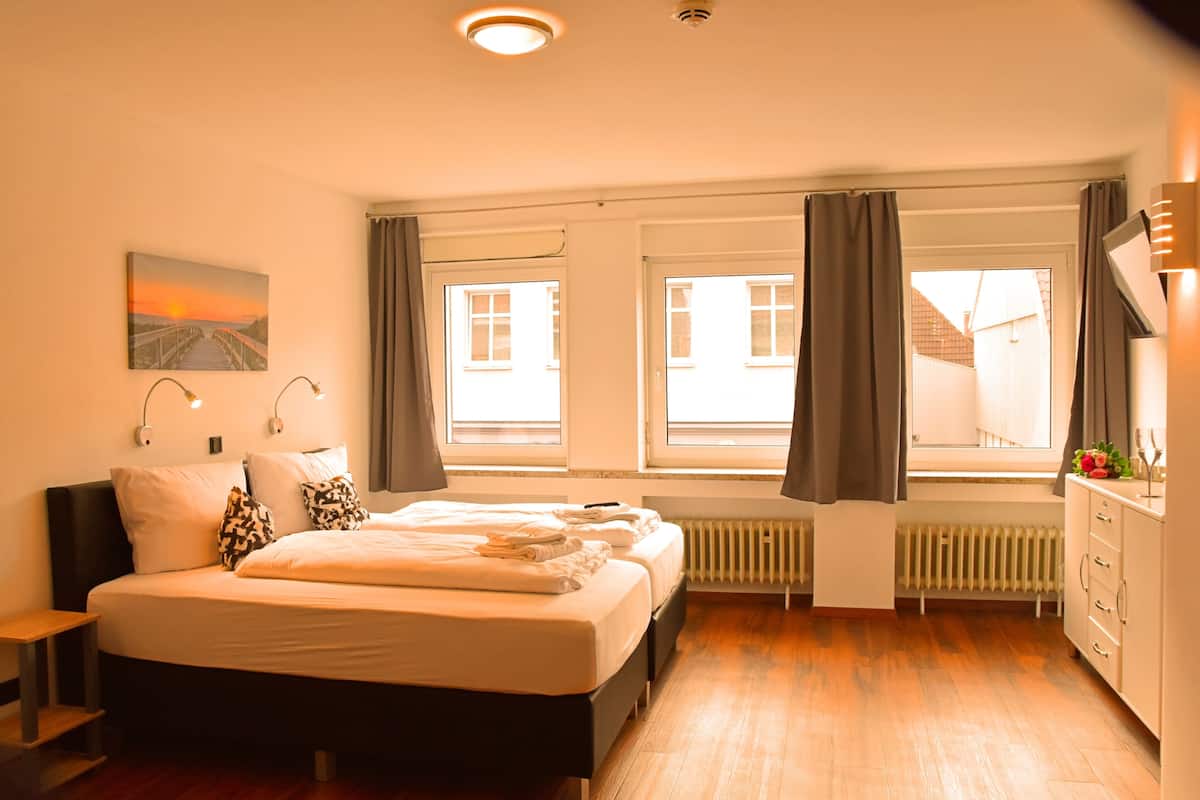 Habitación de hotel con cama, mesitas de noche, radiador, ventana con cortinas y vista a edificios.