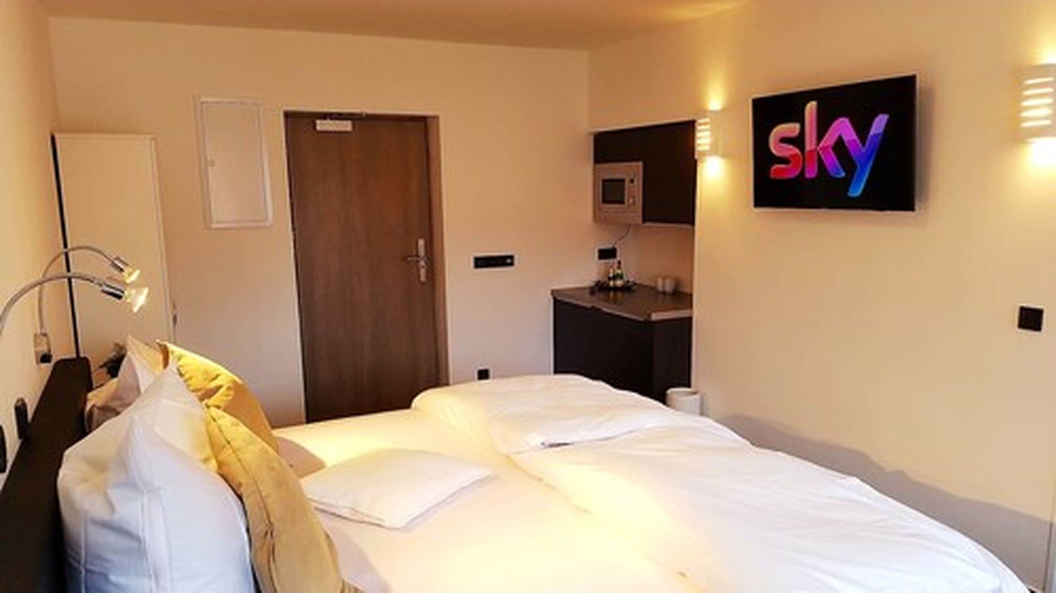 A hotel room with a bed, a desk, a chair, a lamp, a door, and a wall-mounted TV displaying the word "sky".