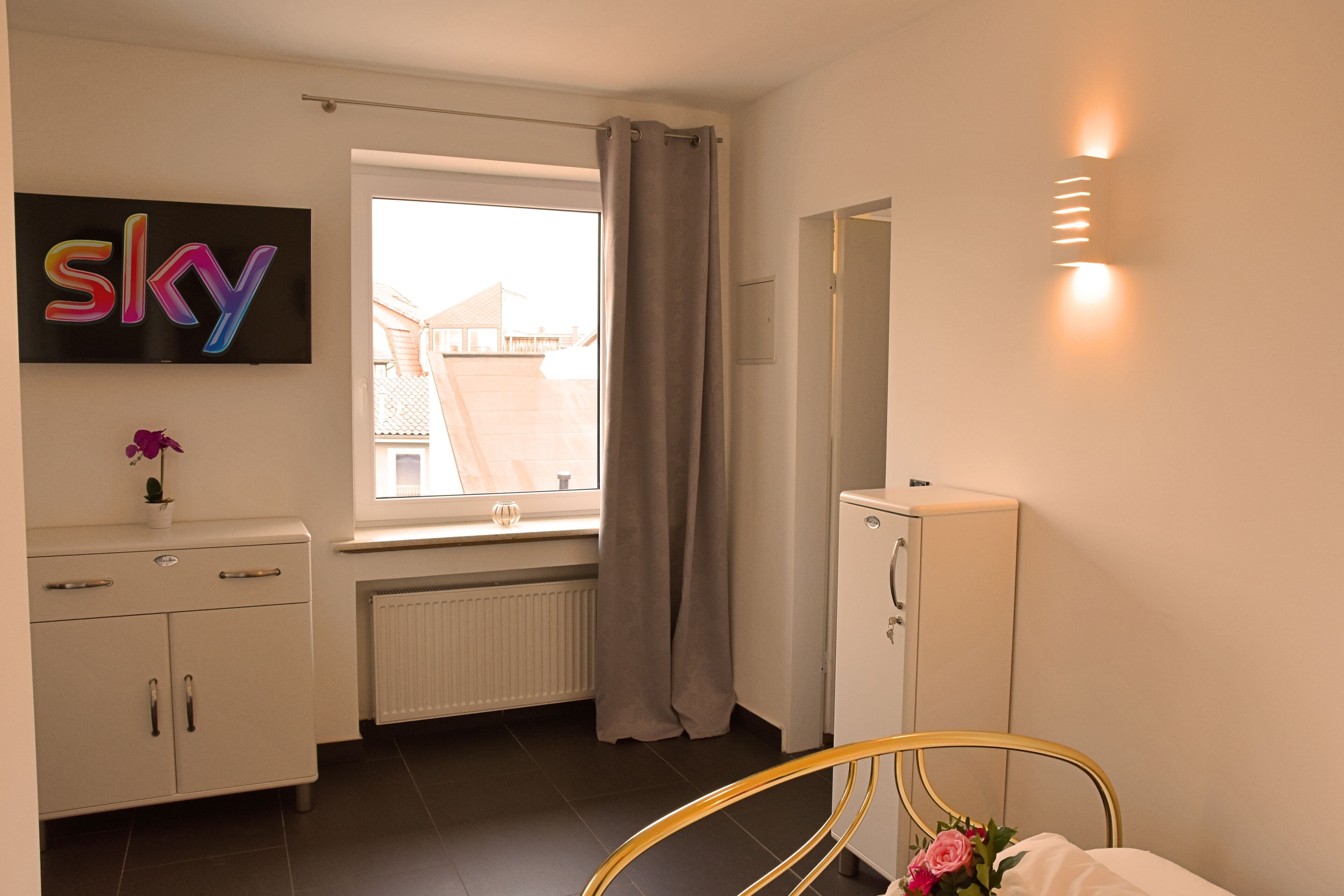A bedroom with a bed, a dresser, a radiator, a window with curtains, a wall-mounted TV, and a mirror.