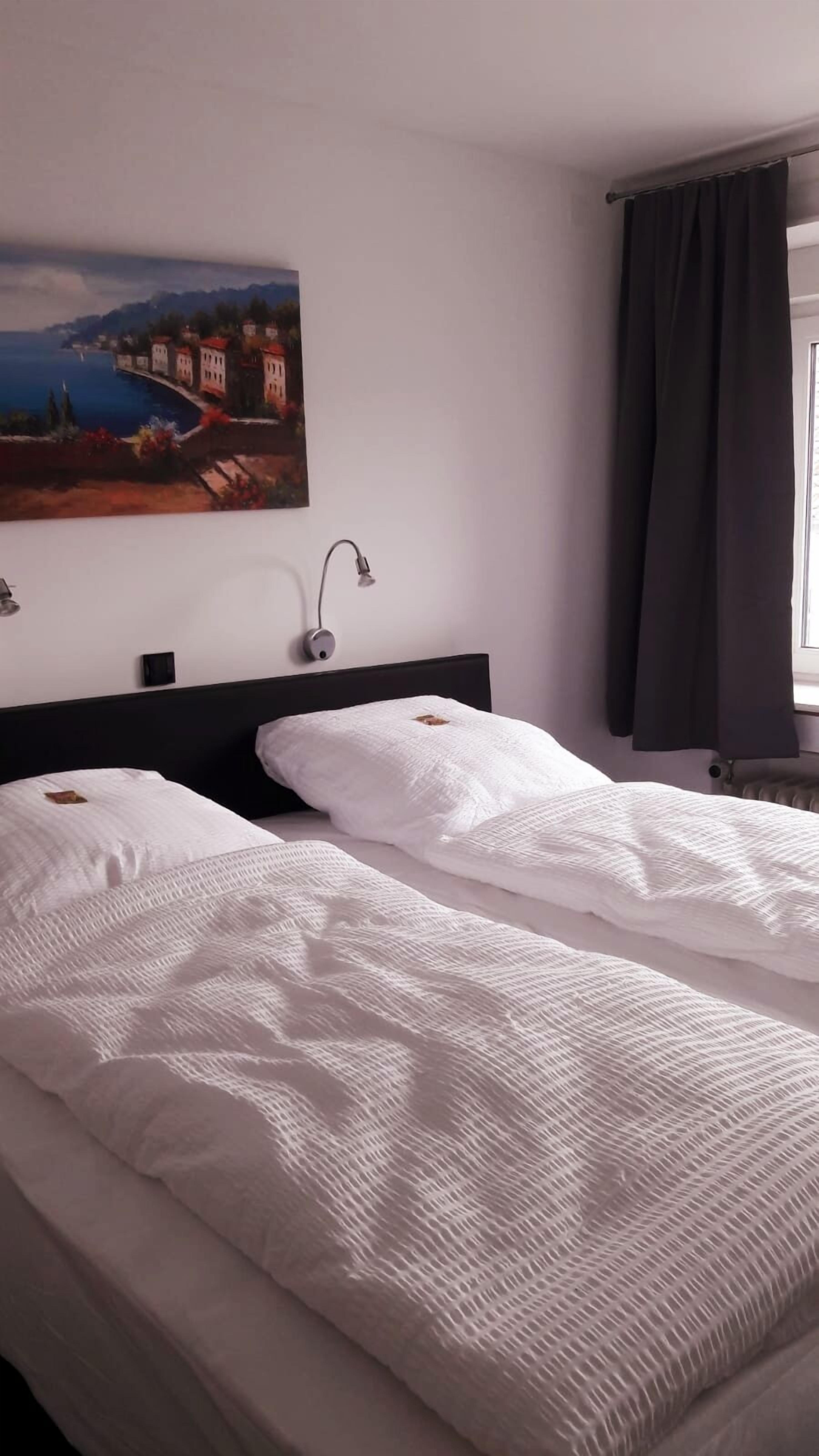Ein Hotelzimmer mit zwei Betten, einem Bild einer Küstenlandschaft, einem Nachttisch und einem Fenster mit Vorhängen.