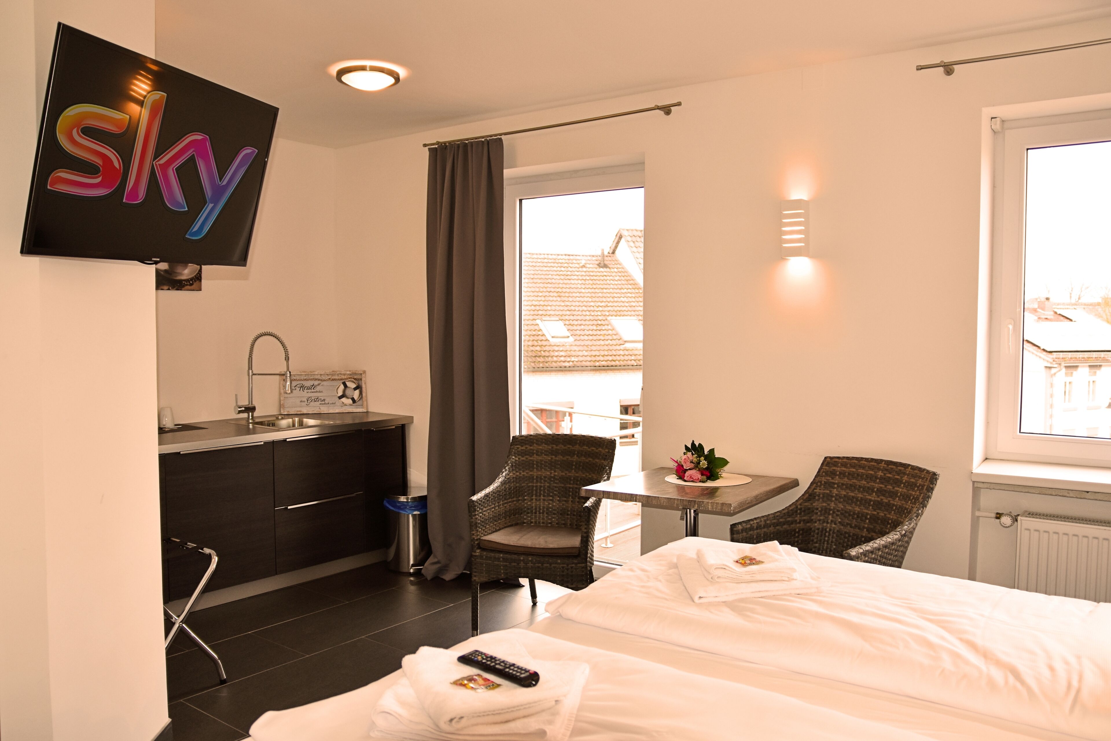 A hotel room with a bed, two chairs, a small table with a flower, a TV mounted on the wall, a window, and a view of buildings outside.