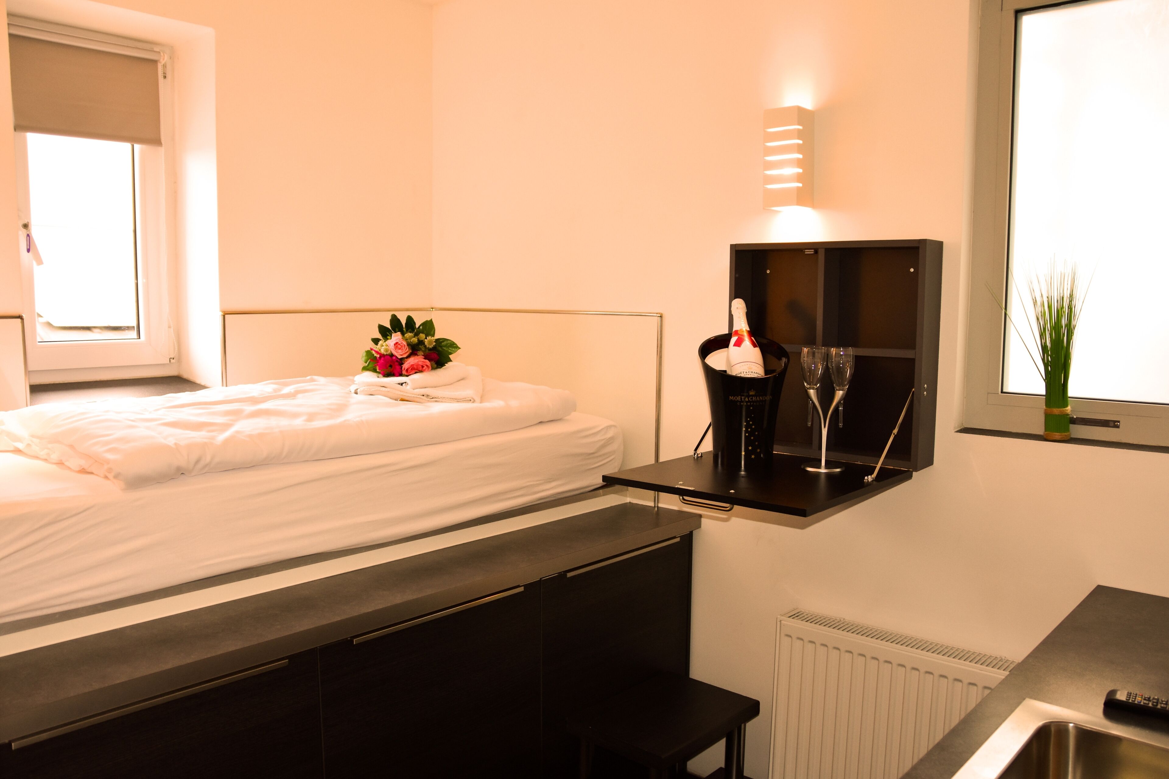 Habitació d'hotel amb un llit, una taula de nit amb una ampolla i gots, una petita estant, un radiador i una finestra amb vistes.