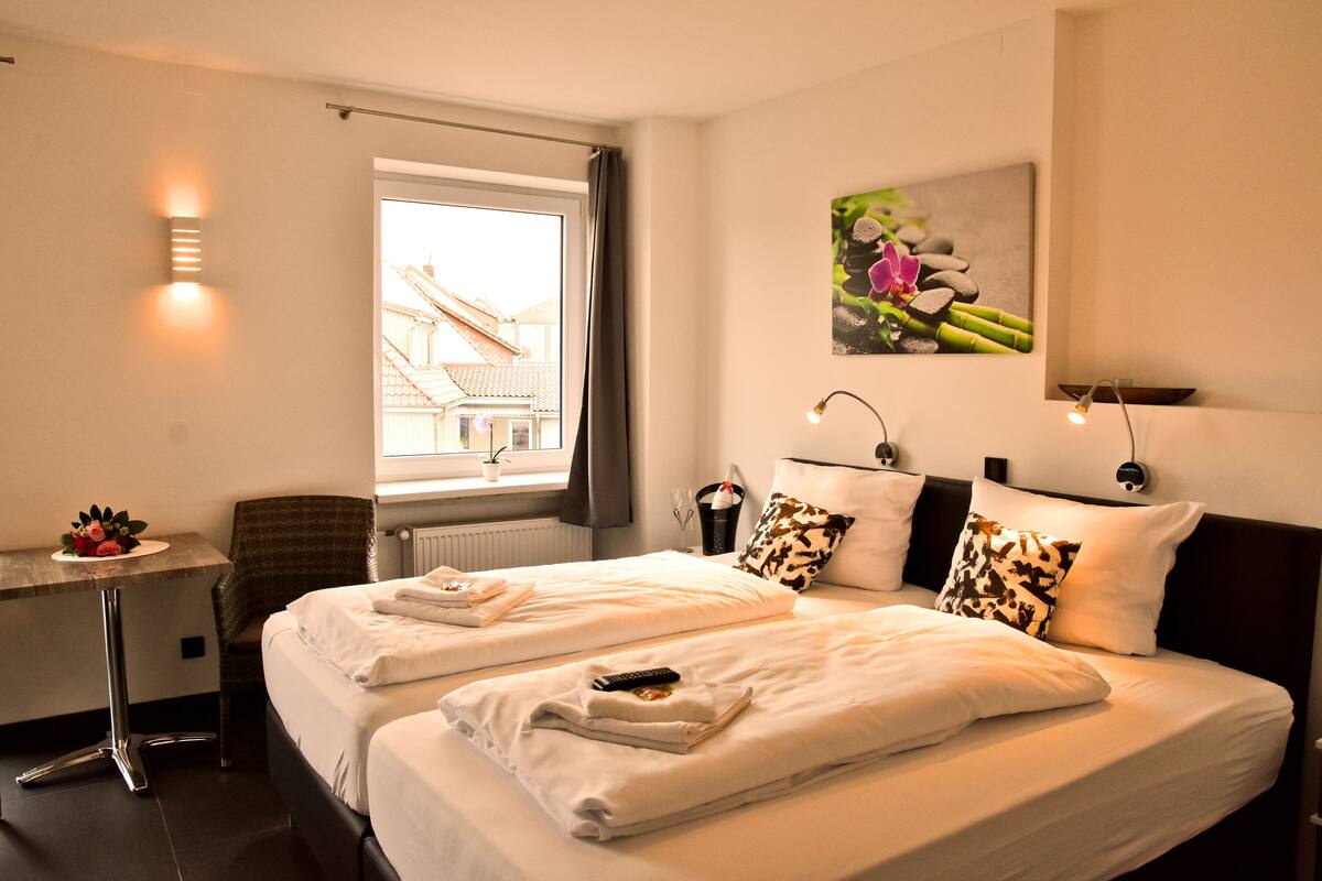 Habitación de hotel con dos camas, un escritorio, una silla, una ventana con vistas a edificios y un cuadro colgado en la pared.