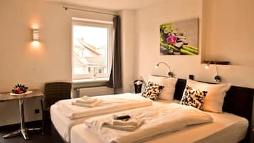 A hotel room with two beds, a desk, a chair, a window with a view of buildings, and a wall-mounted picture.