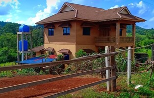 Une maison en bois à deux étages, dotée d’un balcon, entourée d’une végétation luxuriante et de collines.