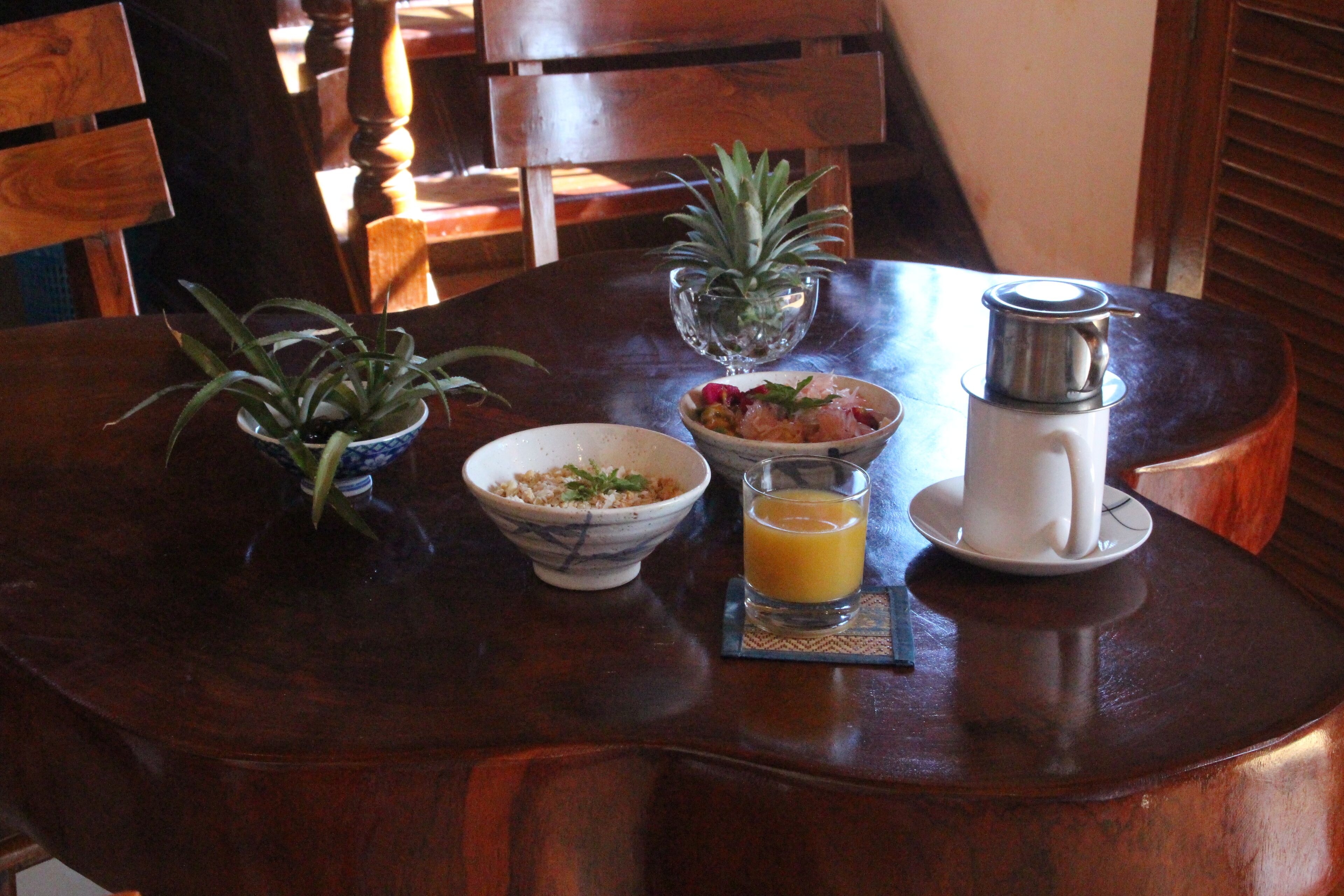 A wooden dining table with a bowl of cereal, a glass of orange juice, a small potted plant, and a coffee maker.