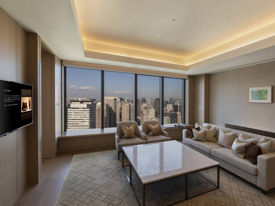 A modern living room with a large window offering a city view, a flat-screen TV, and a sofa with multiple cushions.