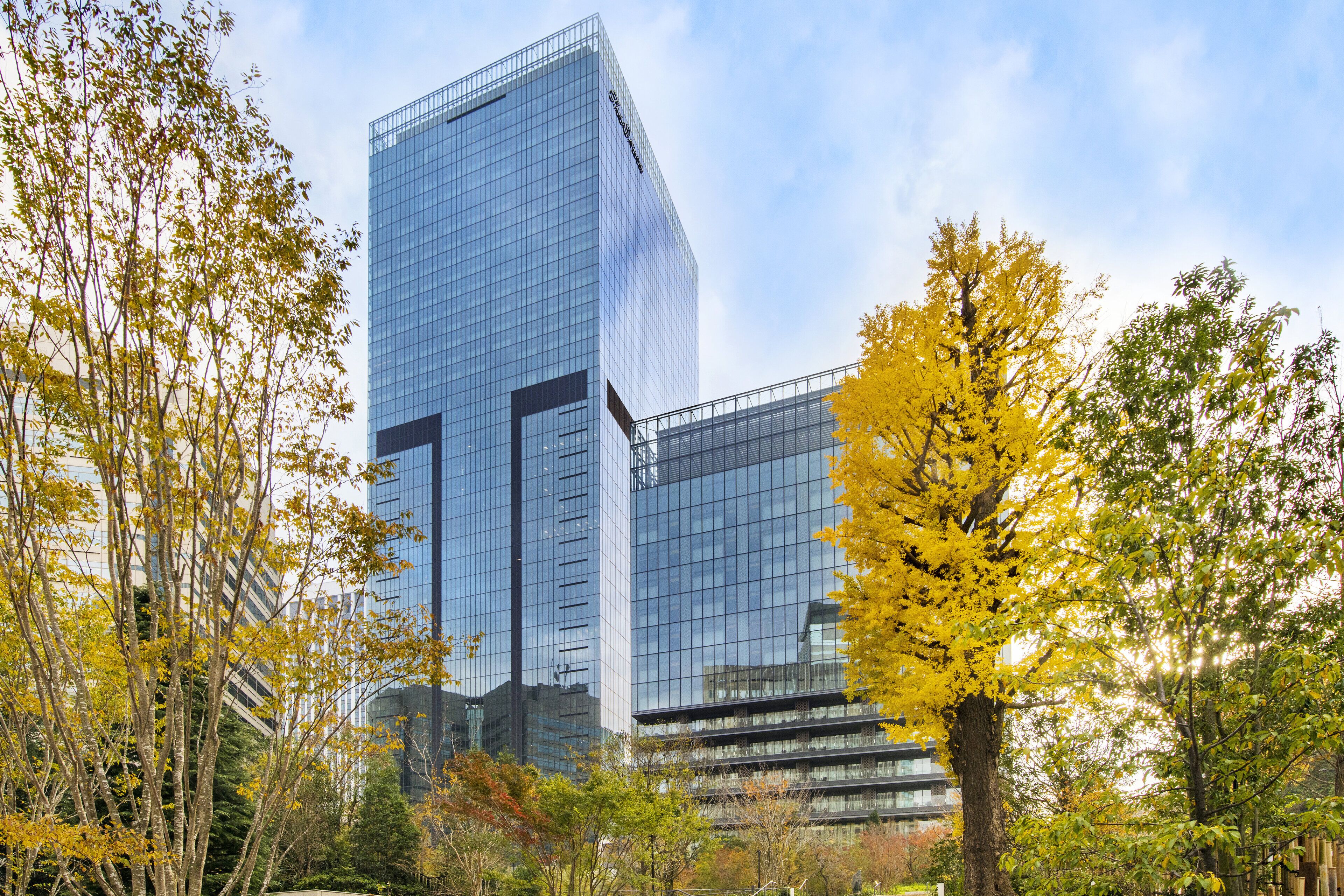 A modern glass building with a reflective facade, surrounded by trees with yellow leaves, indicating an autumn setting.