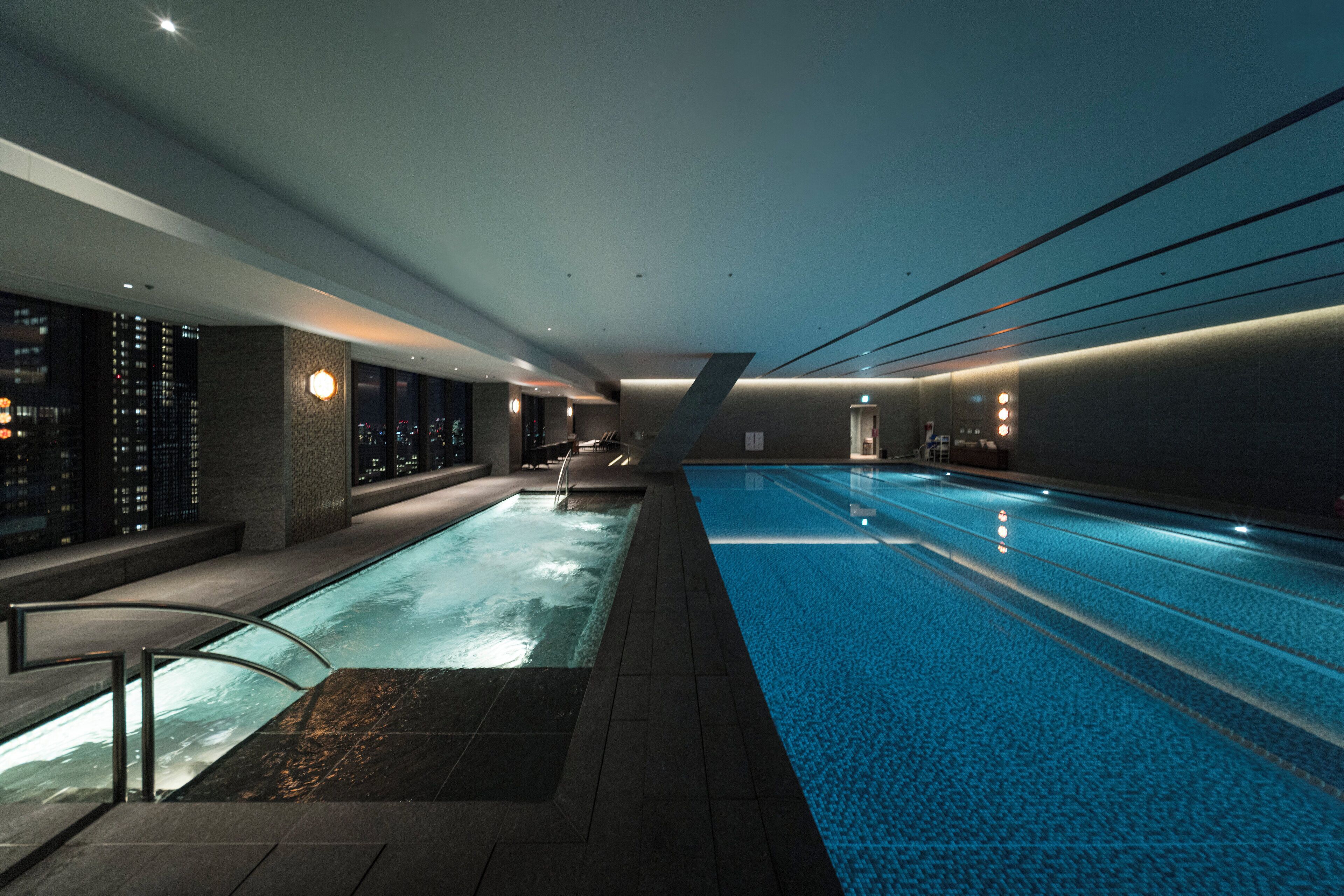 A modern indoor swimming pool with clear water, a separate shallow area, and a seating area along the pool's edge.