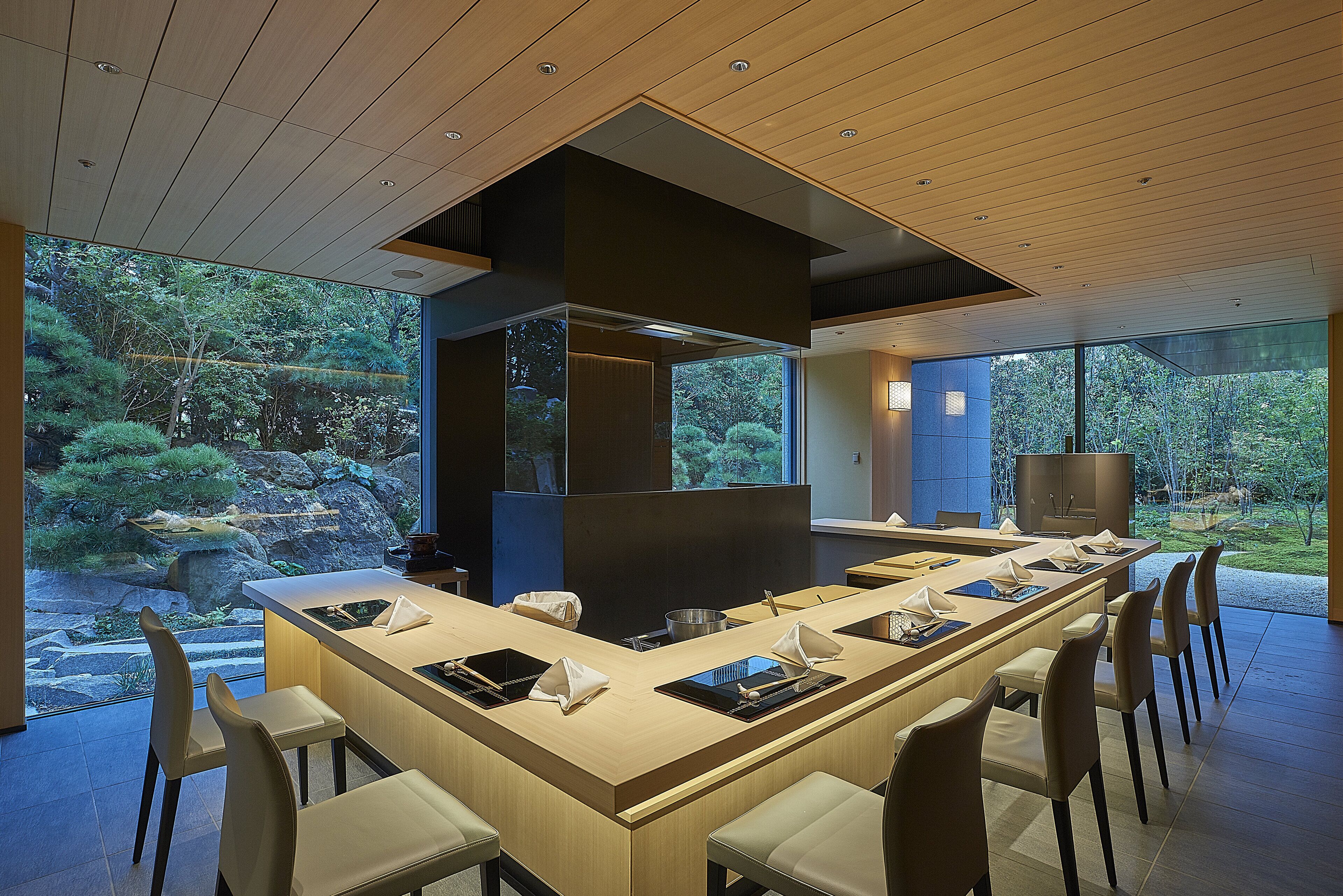 A modern dining area with a long table, multiple chairs, and a view of a garden through large windows.