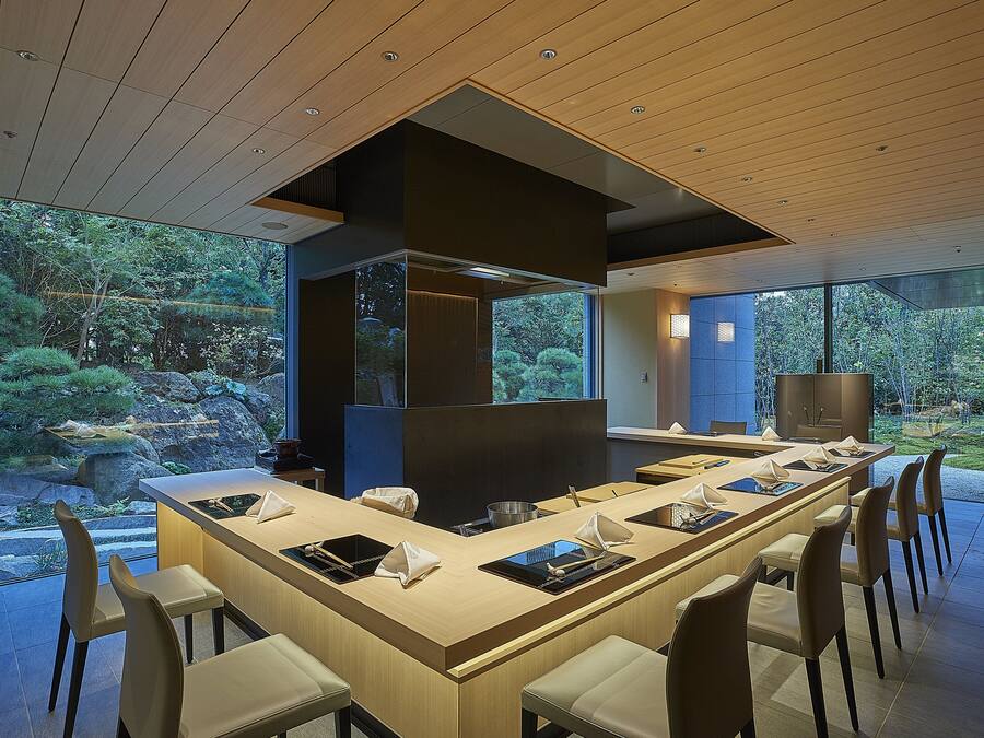 A modern dining area with a long table, multiple chairs, and a view of a garden through large windows.