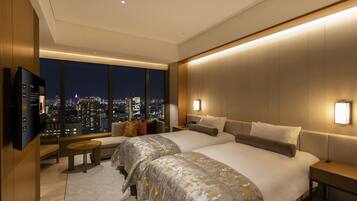 A modern hotel room with a large bed, a TV, a desk, and a view of a cityscape at night.