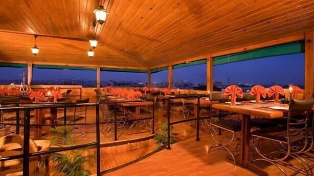 Un restaurante al aire libre con mesas y sillas de madera, toldo verde y vista de la ciudad al anochecer.