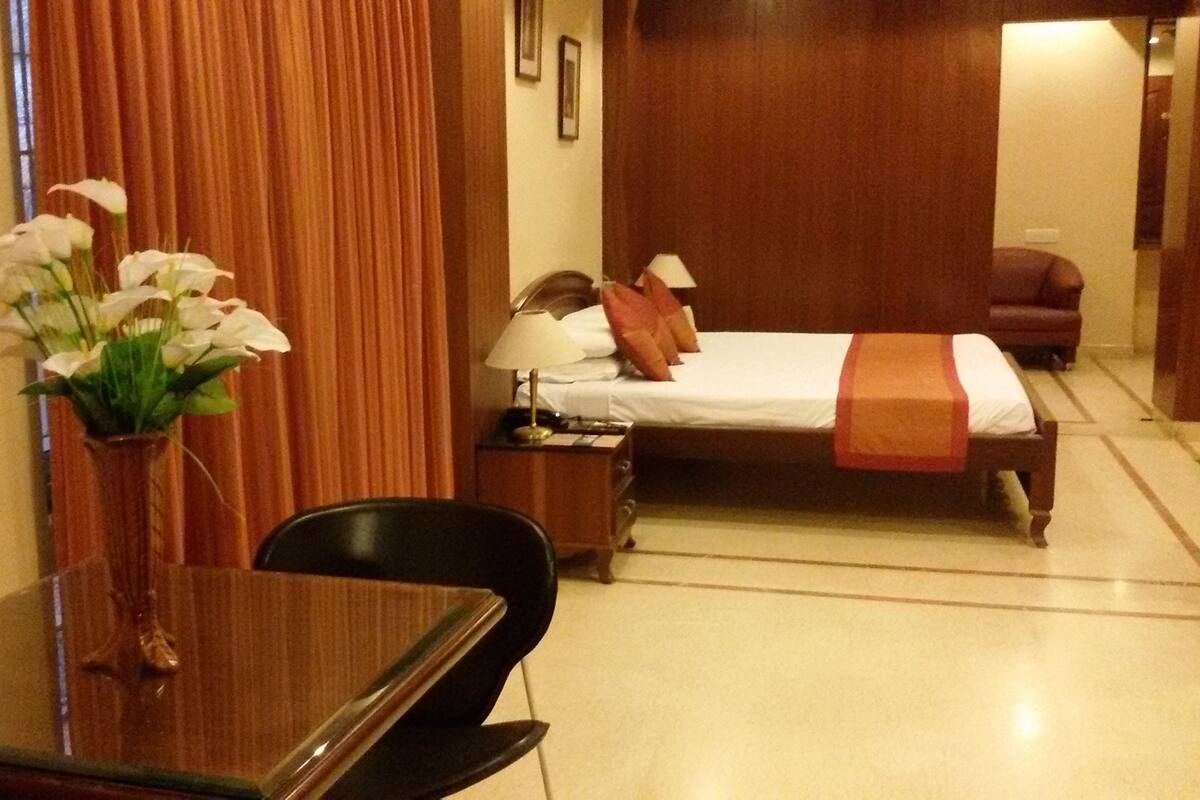 Quarto de hotel com cama, escrivaninha, cadeira, mesa de cabeceira, abajur e um vaso com flores.