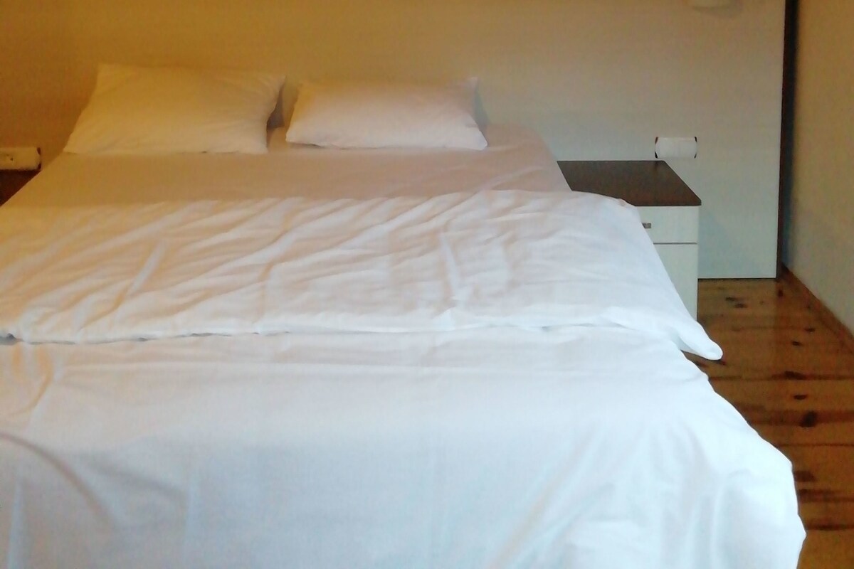 A single-bed room with a wooden ceiling, a white headboard, and a small bedside table with a lamp.