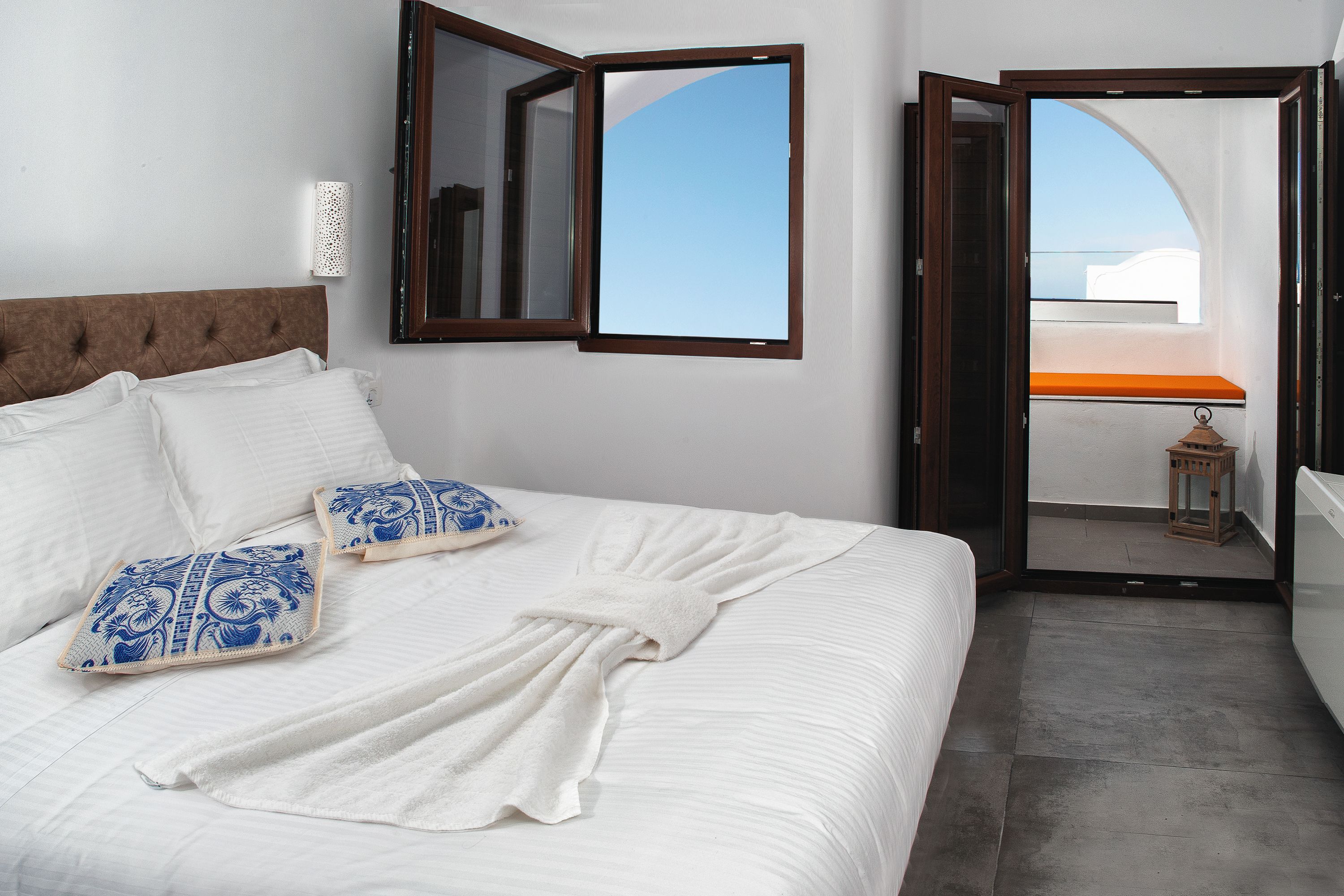 A hotel room with a bed, white bedding, a blue and white pillow, a white robe, a wooden headboard, a wall-mounted mirror, and a balcony with a view.