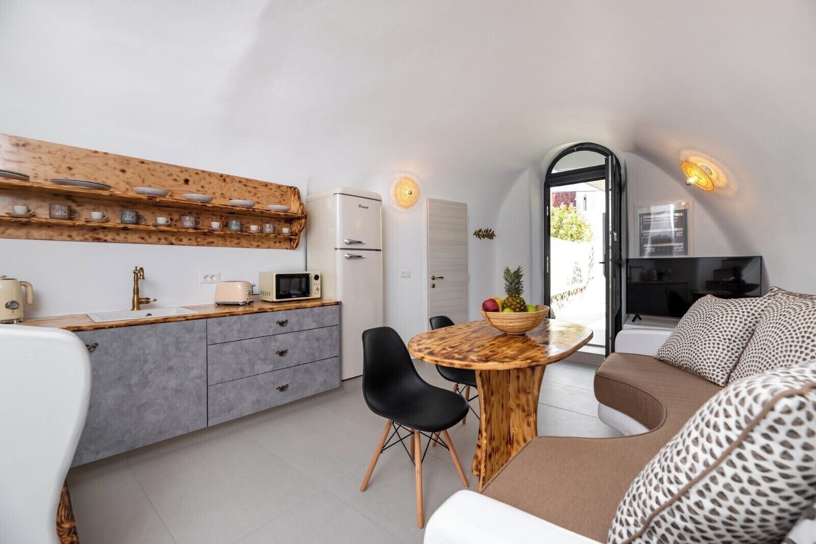 A compact living space with a kitchenette, a dining area with a wooden table and chairs, a sofa with patterned cushions, and a television.