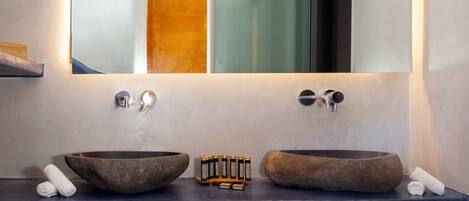 A bathroom with a large mirror above a double sink with stone basins, a towel rack with towels, and a shelf with bottles.