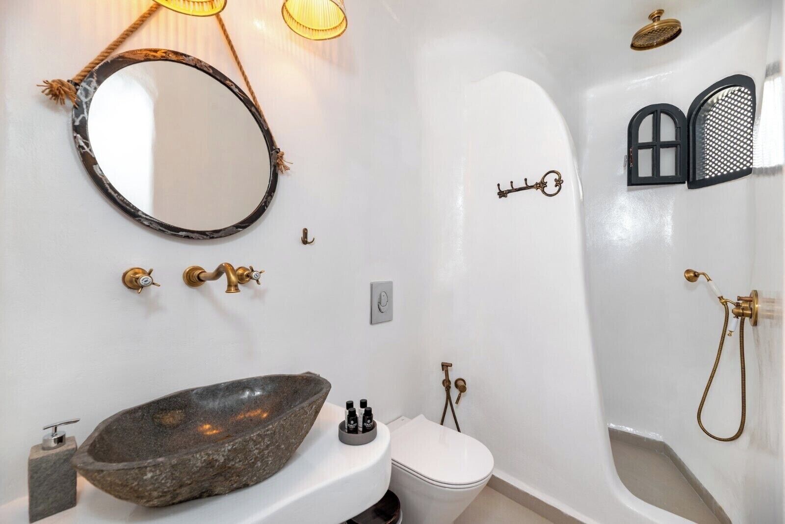Une salle de bain avec un lavabo en pierre, un miroir rond, des toilettes et une douche dont la tête de douche est conçue en forme de trou de serrure.