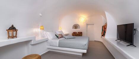 A modern, white-themed bedroom with a bed, bedside lamps, a television, and a small seating area.