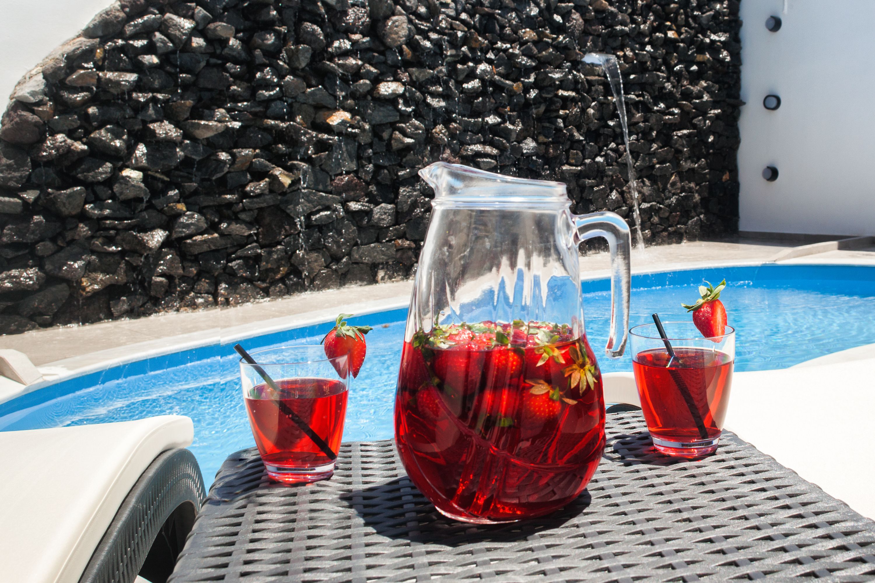 A poolside setting with a pitcher of red drink garnished with strawberries, two glasses of the same drink, and a stone wall in the background.