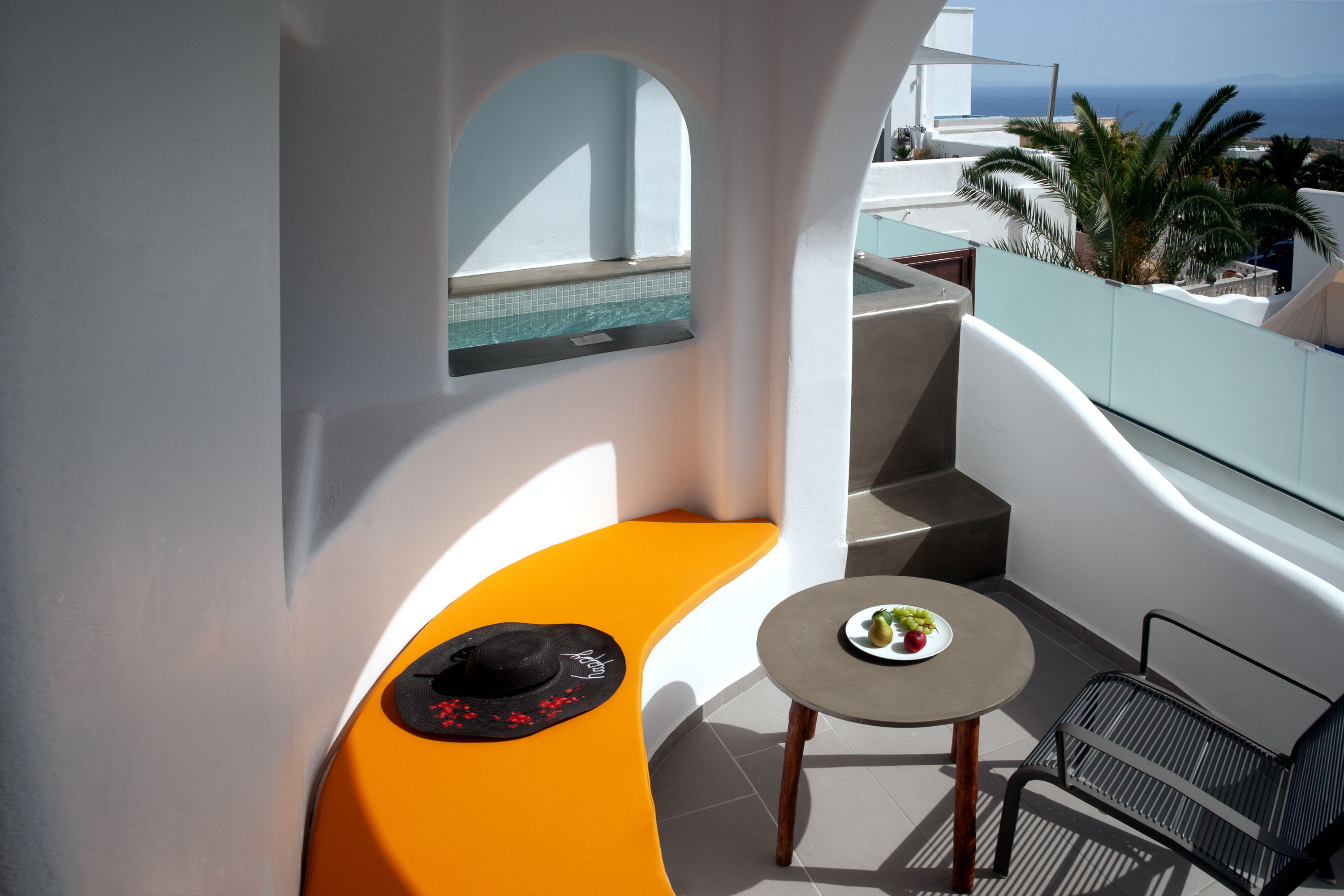 A balcony with a curved orange bench, a small round table with a plate of fruit, and a black hat on the bench.