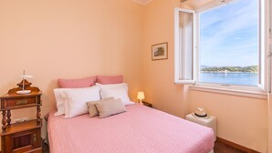 Een slaapkamer met een bed, nachtkastje, lamp, raam met uitzicht op een wateroppervlak en een schilderij aan de muur.