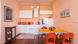 Een moderne keuken met witte kastjes, een inbouwoven en een roestvrijstalen koelkast. Er staat een eettafel met oranje stoelen en een gele placemat.