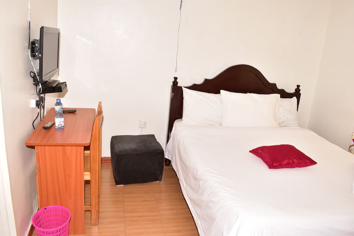 Quarto com escrivaninha de madeira, um banquinho preto, cama com roupa de cama branca e almofada vermelha, e uma televisão montada na parede.