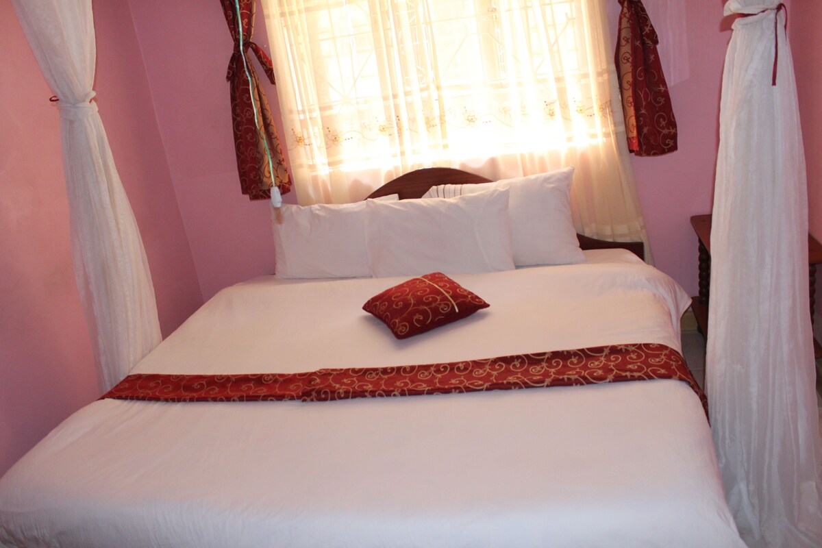 A neatly made bed with white linens and a red decorative pillow, set against a pink wall with sheer curtains.