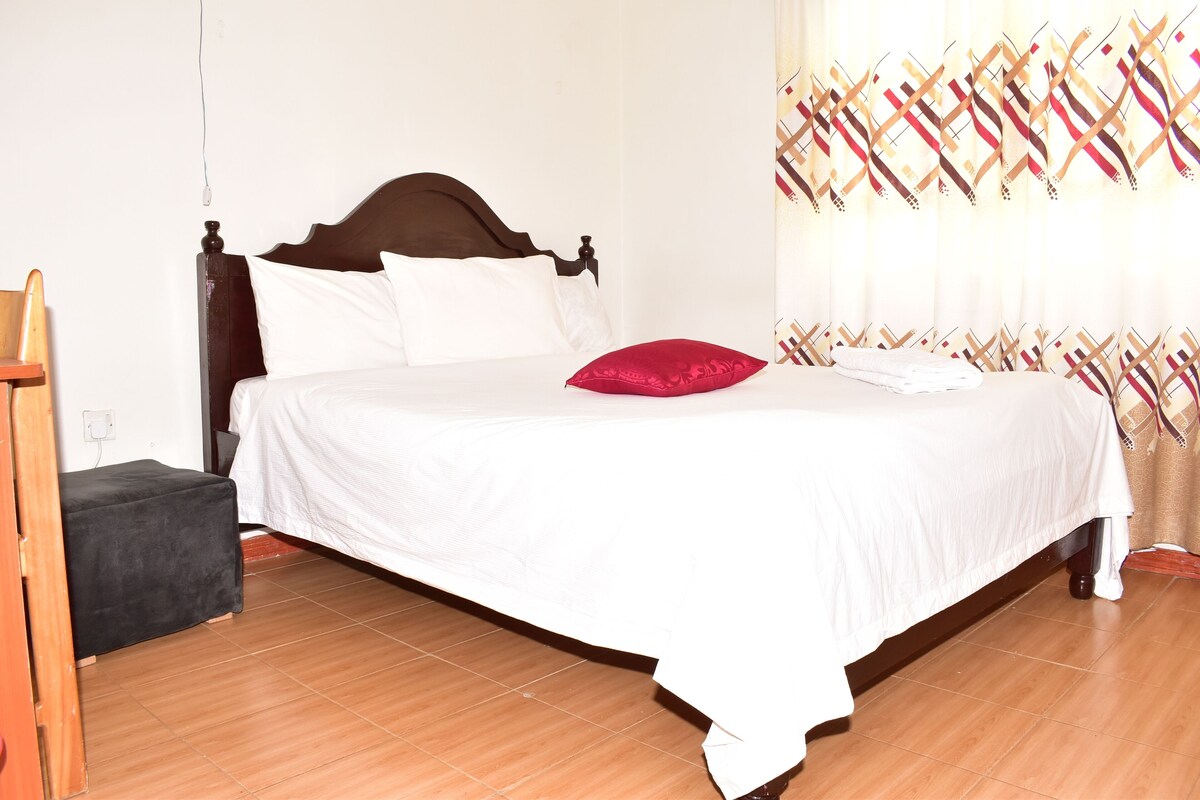 A neatly made bed with white bedding and a red pillow, a wooden headboard, and a small black ottoman.