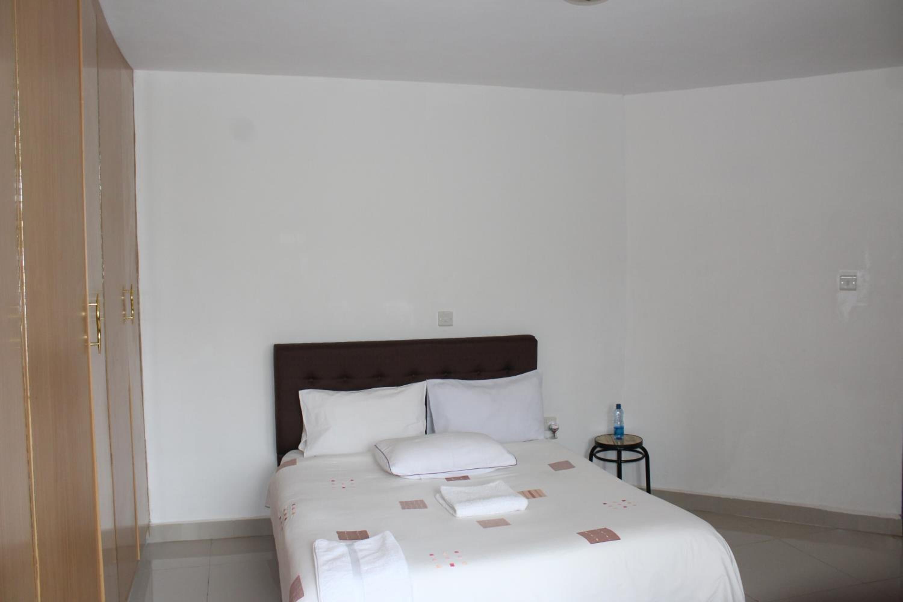 A neatly made bed with white linens and a brown headboard, a small bedside table with a water bottle, and a closed wooden wardrobe.